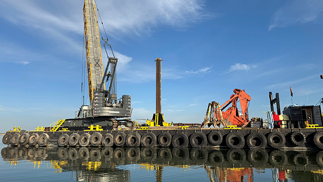 A barge with a tall crane, grapple, and other construction equipment fills the majority of the photograph. The barge has tires lining the outer edge along the waterline. The barge is floating in water that is glassy calm.